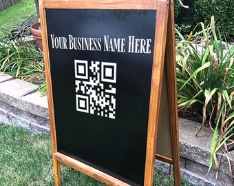 May include: A wooden A-frame sign with a black chalkboard surface. The sign has the text "YOUR BUSINESS NAME HERE" and a black and white QR code printed on it.