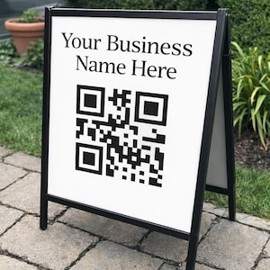May include: A-frame sign with a white panel and black frame. The sign displays the text "Your Business Name Here" above a black and white QR code. The sign is positioned on a brick walkway.