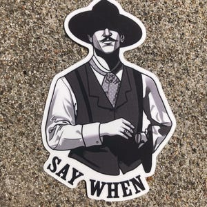 Puede incluir: Adhesivo en blanco y negro que presenta una ilustración estilizada de un hombre con sombrero de vaquero y chaleco, sosteniendo un arma. El texto "SAY WHEN" está en la parte inferior del adhesivo.