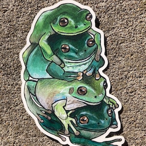 Puede incluir: Una pegatina con una pila de cuatro ranas verdes. Las ranas están dibujadas en un estilo de dibujos animados con ojos grandes y están apiladas una encima de la otra.