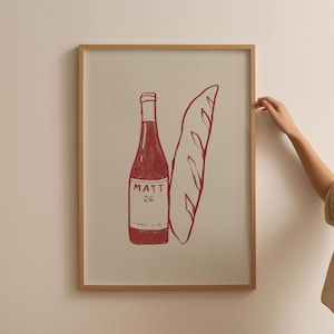 May include: A framed print featuring a red illustration of a wine bottle and a baguette. The wine bottle has the text "MATT 26" on the label. The artwork is in a wooden frame.
