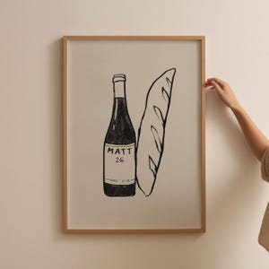 May include: A framed black and white illustration of a wine bottle and a baguette. The wine bottle has the text "MATT 26" on the label. The artwork is in a light brown wooden frame.