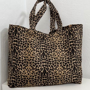 Könnte beinhalten: Eine große Tragetasche mit Leopardenmuster in Braun- und Schwarztönen. Die Tasche hat zwei Griffe und ist aus einem robusten Stoff gefertigt. Die Tasche steht auf einer weißen Oberfläche.