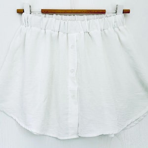 Könnte beinhalten: Ein weißer Rock mit elastischem Bund und Knopfleiste vorne. Der Rock ist aus leichtem Stoff gefertigt und hängt an einem Holzbügel. Der Rock hat einen leicht geschwungenen Saum.