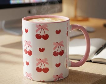 May include: A white ceramic mug with a pink rim and handle, featuring a repeating pattern of red cherries tied with light pink bows and small red hearts. The mug is filled with a hot beverage showing latte art foam on the surface.