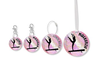 Puede incluir: Un conjunto de cuatro accesorios personalizados con temática de gimnasia. Cada dije redondo presenta una silueta negra de una gimnasta en una barra de equilibrio sobre un fondo de acuarela rosa y dorado. Los nombres están impresos en los dijes.