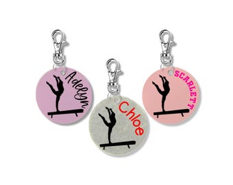 May include: Three pink, gray, and white round keychains with silver clips. Each keychain features a silhouette of a gymnast performing on a balance beam and a name: Adelyn, Chloe, and Scarlett.