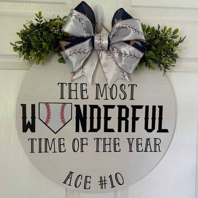 Puede incluir: Un letrero redondo de madera blanca con el texto "The Most Wonderful Time of the Year" y un gr&aacute;fico de b&eacute;isbol. Un lazo de sat&eacute;n blanco con costuras rojas y un patr&oacute;n de b&eacute;isbol azul y blanco est&aacute; unido a la parte superior del letrero. El letrero tambi&eacute;n tiene el texto "Ace #10" en la parte inferior.