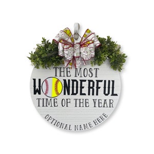 Puede incluir: Un letrero redondo de madera blanca con el texto "The Most Wonderful Time of the Year" y un gr&aacute;fico de softball. El letrero est&aacute; decorado con una cinta roja y blanca con un patr&oacute;n de softball y follaje verde.