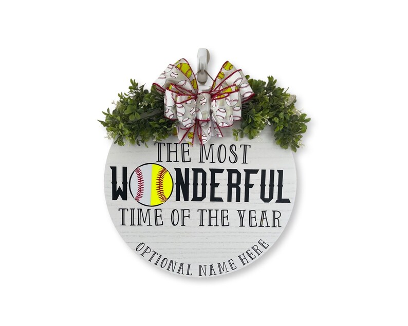 Puede incluir: Un letrero redondo de madera blanca con el texto "The Most Wonderful Time of the Year" y un gr&aacute;fico de softball. El letrero est&aacute; decorado con una cinta roja y blanca con un patr&oacute;n de softball y follaje verde.