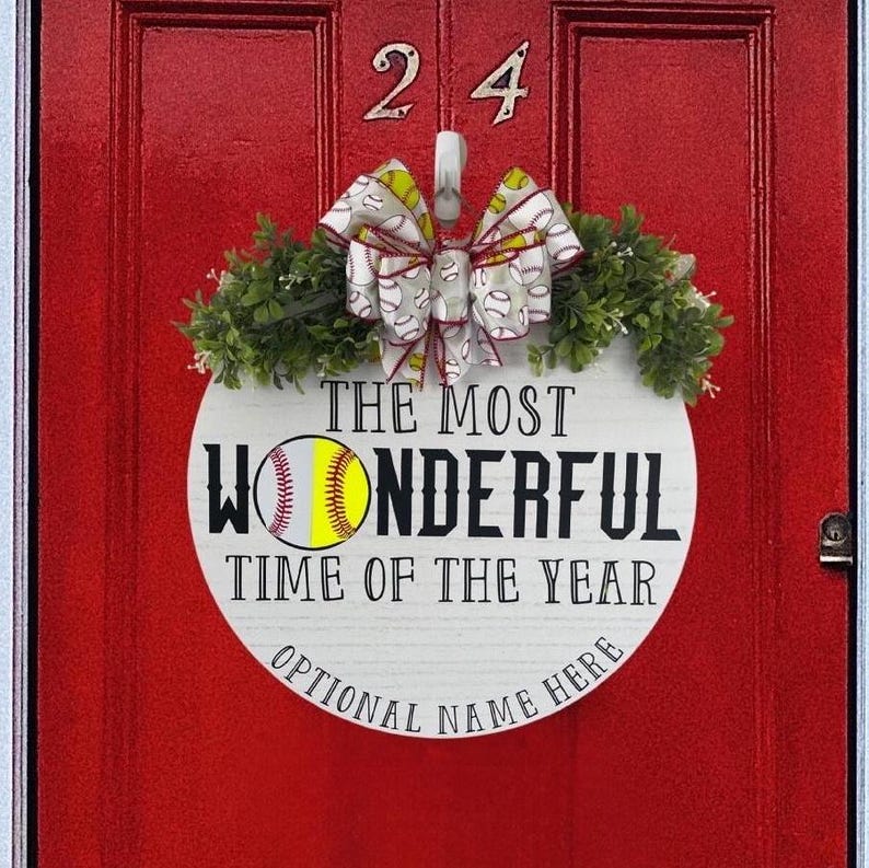 Puede incluir: Un letrero redondo de madera blanca con el texto "The Most Wonderful Time of the Year" y un gr&aacute;fico de softbol. El letrero est&aacute; decorado con un lazo blanco y amarillo y follaje verde.
