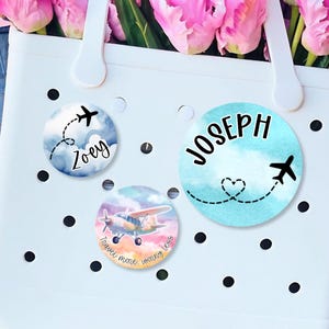May include: Three round, blue and white, watercolor-style stickers with airplane designs. One sticker says "Zoey" with a small airplane and a dotted line. Another sticker says "Joseph" with a heart-shaped dotted line and a small airplane. The third sticker has a small airplane and the text "Travel more, worry less".