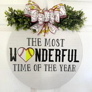 Puede incluir: Un letrero redondo de madera blanca con el texto "The Most Wonderful Time of the Year" en letras negras. Una pelota de softball con costuras rojas tiene la forma de un coraz&oacute;n en el medio de la palabra "Wonderful". Un lazo blanco y amarillo con un patr&oacute;n de pelota de softball est&aacute; en la parte superior del letrero.