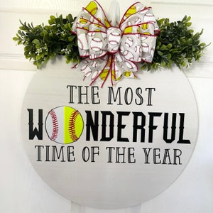 Puede incluir: Un letrero redondo de madera blanca con el texto "The Most Wonderful Time of the Year" en letras negras. Una pelota de softbol se muestra a la izquierda del texto. Un lazo blanco y amarillo con dise&ntilde;os de pelotas de softbol est&aacute; unido a la parte superior del letrero.