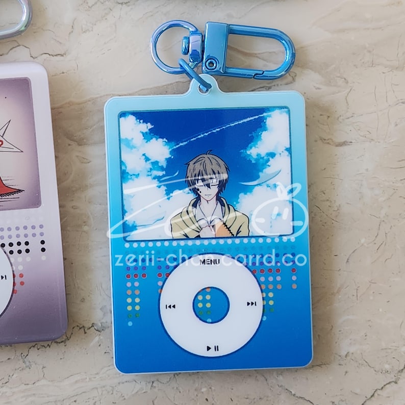 May include: A blue acrylic keychain featuring an anime character design. The keychain has a blue frame with a sky background and a character illustration. The text "zerii-charrd.co" is visible. Includes a blue metal clasp.