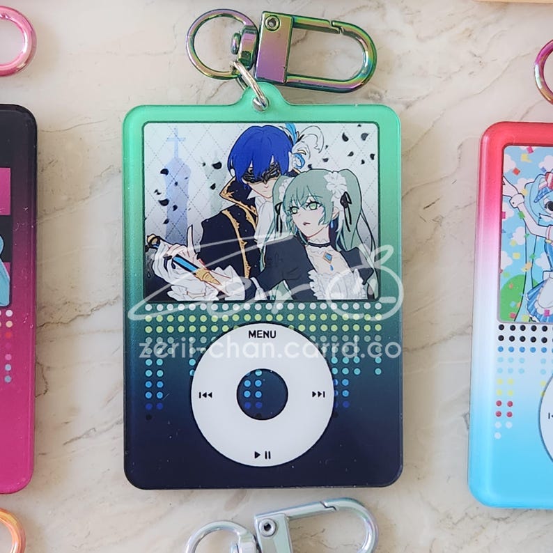 May include: A teal and navy blue keychain featuring an anime-style illustration of two characters. The keychain has a gradient design and a silver-colored clasp. The text "MENU" and "zeri-chan.co.go" are visible.