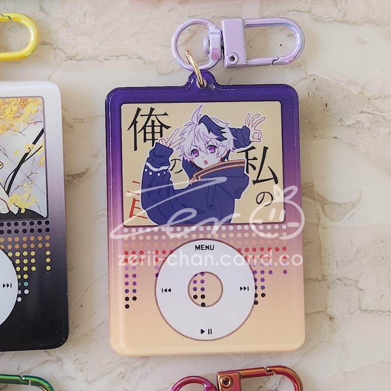 May include: A purple and gold keychain shaped like a music player, featuring a cartoon character illustration. The keychain has a silver clasp and the text "MENU" and "zorii-chan.carrd.co". The illustration includes Japanese text.