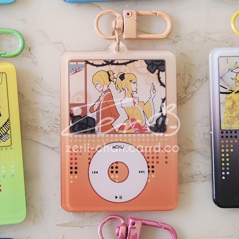 May include: Orange acrylic keychain with a gold-colored clasp. The keychain features a design resembling a music player with a screen displaying two anime characters. The text "zerii-chan.carrd.co" and "MENU" are visible on the keychain.