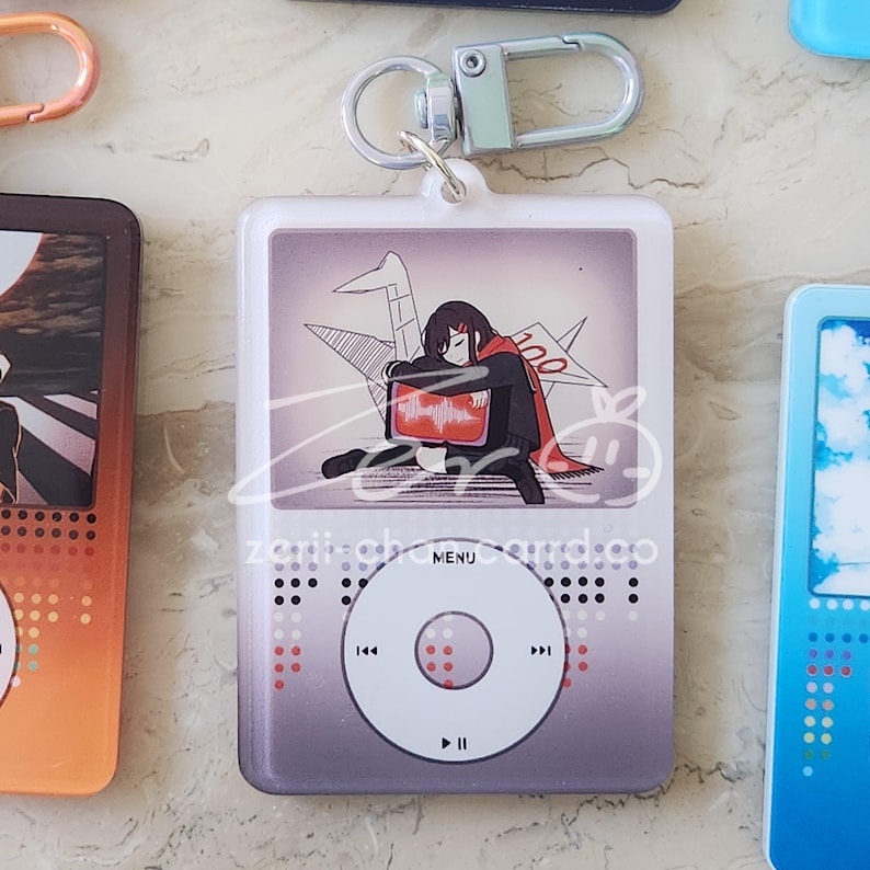 May include: A white and gray acrylic keychain shaped like a music player, featuring a cartoon illustration of a person with a red cape. The keychain has a silver clasp and the word "MENU" is printed on the front.