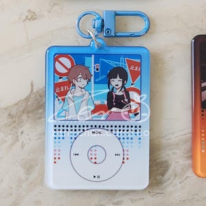 May include: A blue keychain featuring a cartoon design of two anime characters and Japanese text. The keychain is shaped like a music player with a blue frame and a silver-colored clip.