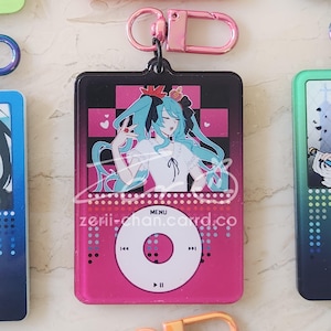 May include: A pink and black acrylic keychain featuring a cartoon character with long turquoise hair. The design resembles a music player with a menu button and control icons. The text "zerii-chan.card.co" is visible.
