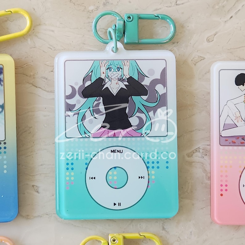 May include: A teal and white keychain featuring a cartoon character with teal hair and a black top. The keychain has a teal and white iPod design with the text "zerii-chan.card.co".