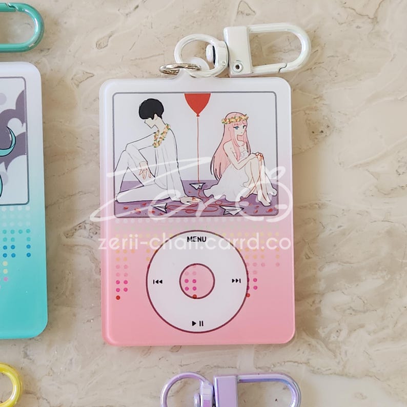 May include: Pink and white acrylic keychain featuring a cartoon illustration of two figures with a red balloon. The keychain has a design resembling a music player, with a circular control pad and a silver-colored clasp.