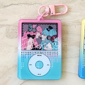 May include: A pink and blue keychain featuring a cartoon illustration of two anime characters. The keychain has a pink border and a blue to pink gradient background. The design resembles a music player with a menu button and control icons.