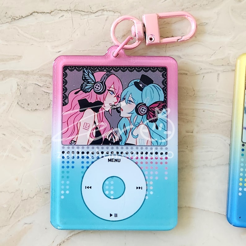 May include: A pink and blue keychain featuring a cartoon illustration of two anime characters. The keychain has a pink border and a blue to pink gradient background. The design resembles a music player with a menu button and control icons.