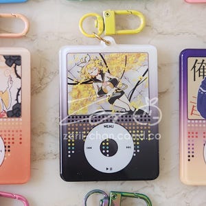 May include: A white and black keychain featuring a cartoon character design. The design resembles a music player with a screen displaying a yellow-toned illustration. The keychain has a yellow clasp and the text "MENU".