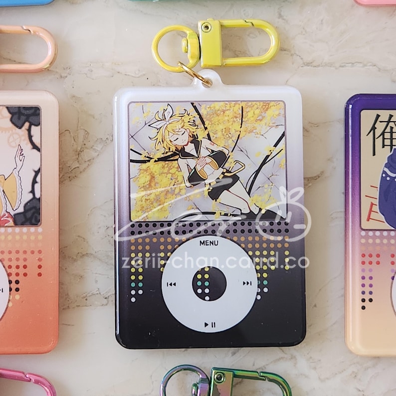 May include: A white and black keychain featuring a cartoon character design. The design resembles a music player with a screen displaying a yellow-toned illustration. The keychain has a yellow clasp and the text "MENU".