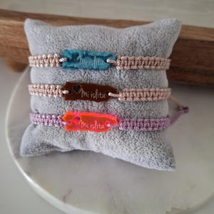 May include: Three macrame bracelets with rectangular tags that read "Mi islita". The tags are blue, brown, and orange. The bracelets are beige, beige, and purple. The bracelets are displayed on a gray pillow.