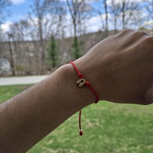 May include: Red string bracelet with a gold-colored letter "A" charm. The bracelet is tied with a sliding knot for adjustable sizing. The background shows a blurred view of trees and a blue sky.