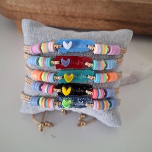 May include: A collection of colorful beaded bracelets with the text "Mi islita" on each. The bracelets feature a variety of pastel-colored beads and small heart-shaped charms. The bracelets are displayed on a gray pillow.