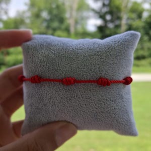 May include: A bright red cord bracelet featuring four evenly spaced decorative knots. The thin, vibrant red string is constructed from a smooth, durable material and is designed to be worn around the wrist as a simple, minimalist accessory.