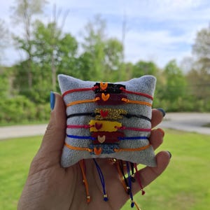 May include: A collection of colorful friendship bracelets with heart-shaped charms. The bracelets are displayed on a small gray pillow and feature various colors like red, orange, black, pink, yellow, blue, and glittery accents.