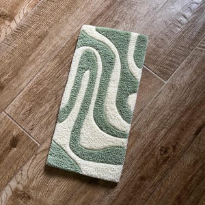 May include: A rectangular rug with a tufted design in shades of green and off-white. The pattern features flowing, curved lines, creating a soft, textured appearance. The rug is placed on a wooden floor.