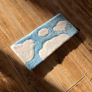 May include: A rectangular rug with a cloud design. The rug has a light blue background with white cloud shapes. The rug is made of a soft, textured material. The rug is placed on a wooden floor.