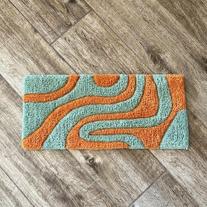 May include: A rectangular rug with a retro design. The rug features a teal background with orange, wavy lines. The rug is placed on a wooden floor with a herringbone pattern. The rug is approximately 24 inches long and 12 inches wide.