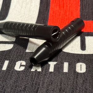 May include: Two black metal darts with a textured grip and a silver tip. The darts are lying on a black and white fabric with the word "RICATION" printed on it.