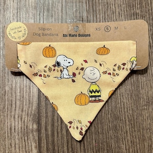 May include: A dog bandana with a fall theme. The bandana features Snoopy and Charlie Brown with pumpkins and falling leaves on a light yellow background. The bandana is attached to a cardboard tag with the text "handmade with love".