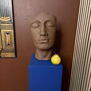 Op de afbeelding: Een bruine sculptuur van een gezicht met gesloten ogen, geplaatst op een blauwe sokkel. Een gele peer rust op de sokkel. De sculptuur staat voor een bruine muur en een decoratief paneel.