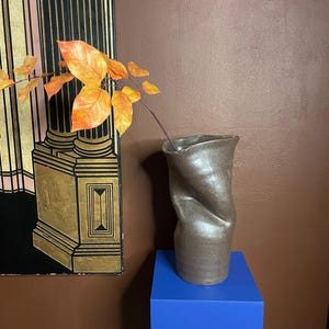 Op de afbeelding: Een bruine keramische vaas met een gedraaid ontwerp, met een tak met oranje bladeren. De vaas staat op een blauwe kubus, met een decoratief scherm op de achtergrond.