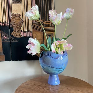 Op de afbeelding: Een blauwe keramische vaas met een bolvormig lichaam en een voetstukbasis bevat verschillende witte en roze tulpen. De vaas heeft een textuur met donkerblauwe accenten. De bloemen hebben gekreukelde bloemblaadjes en groene stelen.