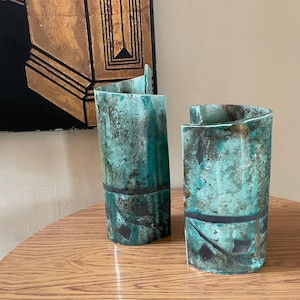 Könnte beinhalten: Zwei türkisfarbene Keramikvasen mit strukturierter Oberfläche und einem dunklen Band in der Mitte. Die Vasen haben ein einzigartiges, geschwungenes Design. Sie stehen auf einem Holztisch.