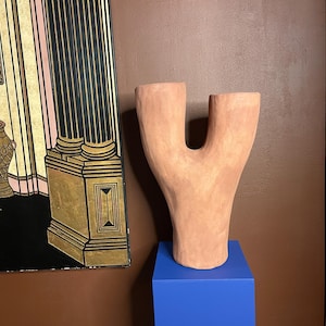 Könnte beinhalten: Eine terrakottafarbene, Y-förmige Keramikvase steht auf einem leuchtend blauen quadratischen Sockel. Die Vase hat eine glatte, organische Form. Der Hintergrund umfasst einen dekorativen Paravent mit goldenen und schwarzen Akzenten.