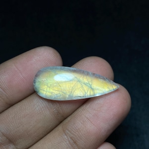 May include: A teardrop-shaped, polished gemstone with a translucent, iridescent quality. The stone displays a mix of yellow, gold, and blue hues, with delicate internal veining. The gem is held in a hand against a dark background.