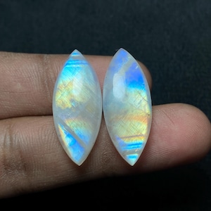 Könnte beinhalten: Zwei marquiseförmige Mondstein-Cabochons. Die Steine zeigen eine perlmuttartige weiße Basis mit schillernden Blitzen in Blau, Gelb und Orange. Die Edelsteine sind poliert und glatt, mit hohem Glanz. Der Hintergrund ist dunkel.