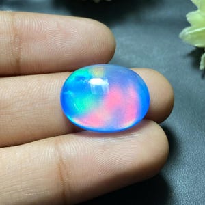 Puede incluir: Un cabujón de ópalo redondo e iridiscente con un juego de colores azul, rosa y verde. El ópalo es liso y pulido.