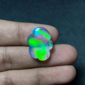 May include: A colorful, iridescent opal gemstone with a unique, flower-like shape. The stone displays vibrant green, blue, and purple hues, with a polished surface. The gemstone is held in a hand, showcasing its natural beauty.